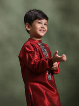 A young boy smiling and gesturing while wearing a shiny red traditional long-sleeve kurta with silver embroidery on the chest, set against a dark olive green studio background.