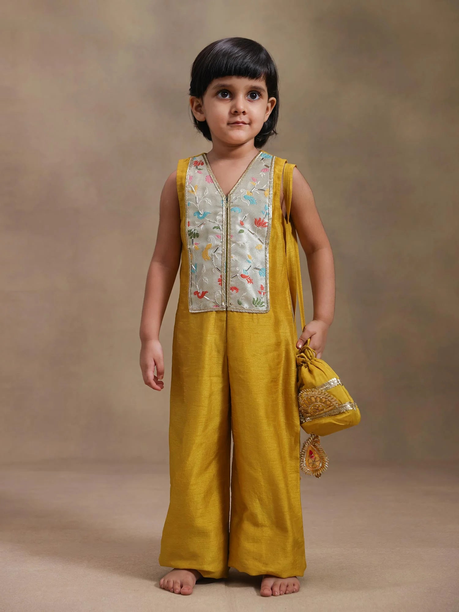 Girl wearing Storytellers Junior Sona Mustard Embroidered Jumpsuit with floral embroidery and potli bag