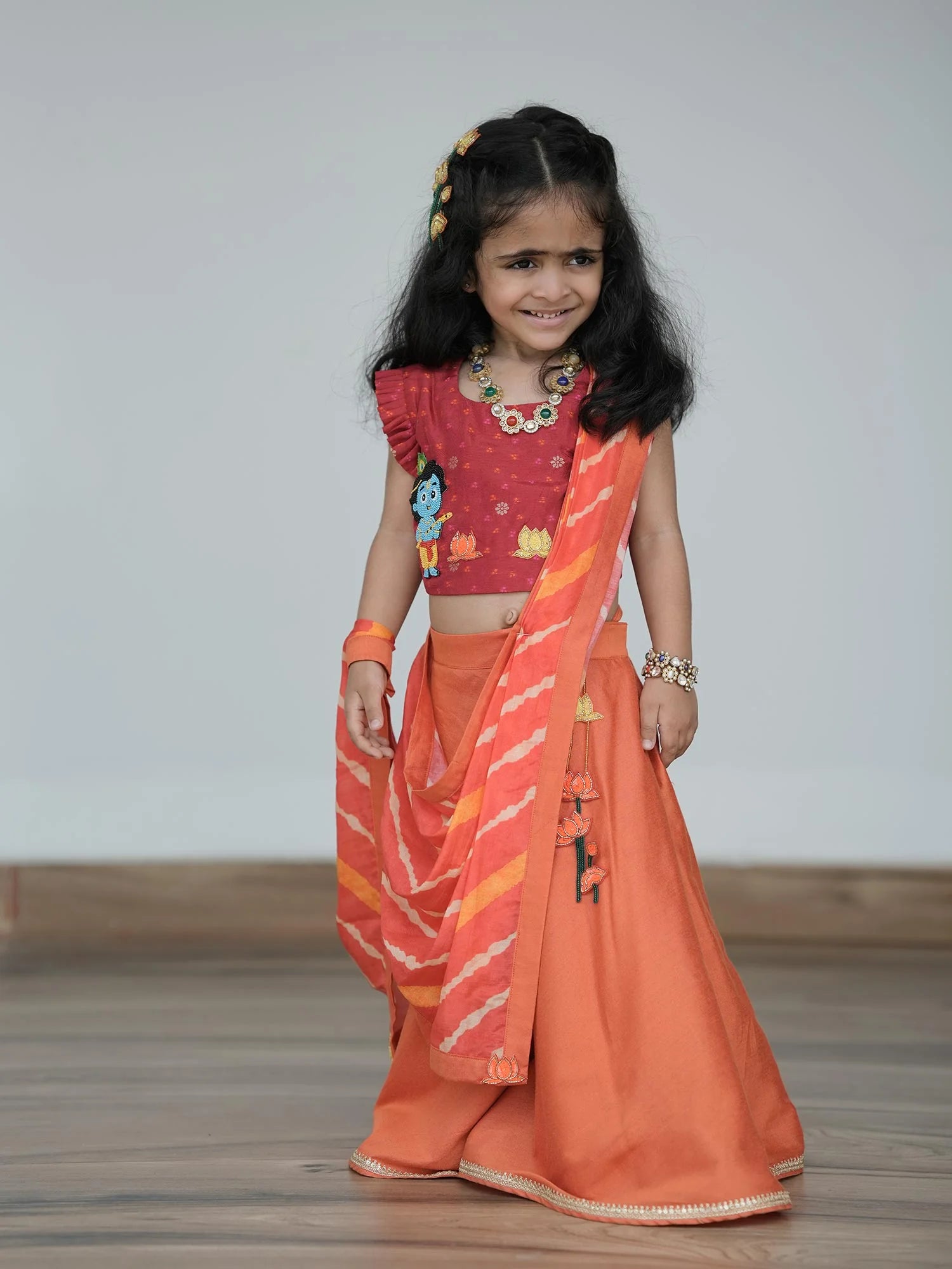 Girl wearing Storytellers Junior Radha Lehenga Choli Set in red and orange for Janmashtami